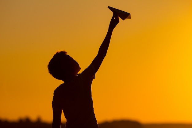 Little boy holds a paper airplane in his hand at sunset. A child raised his hand up to sky and plays with origami in the evening in the street