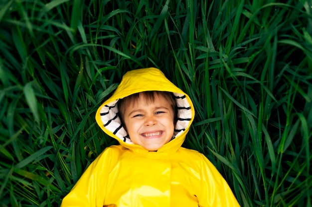 Photo a little boy lies in the thick green grass in a yellow waterproof jacket and smiles.