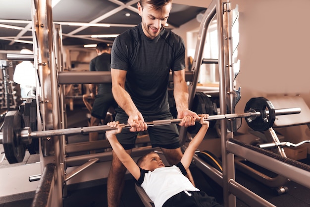 little boy lifts the barbell and his father helps him.