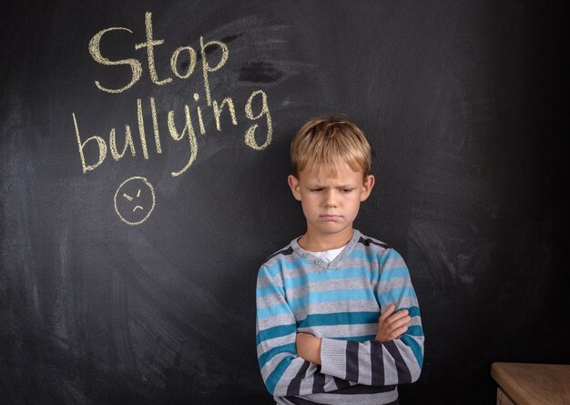 Little boy near chalkboard with words "Stop bullying" indoors