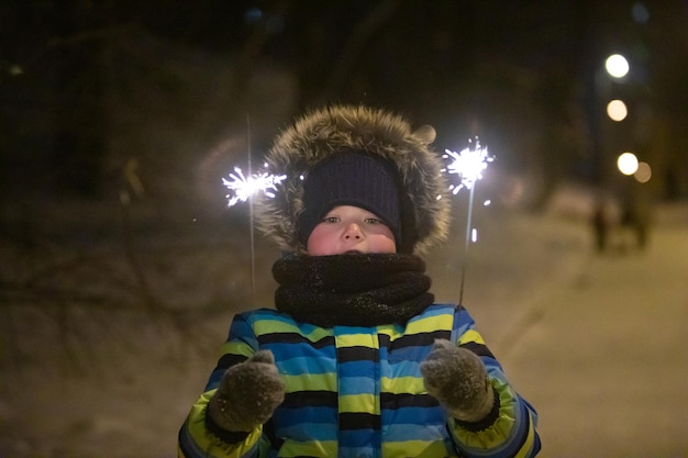 Little boy at night winter city park in warm coat with sparkler