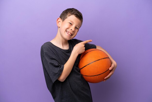 Little boy playing basketball isolated on purple background pointing finger to the side