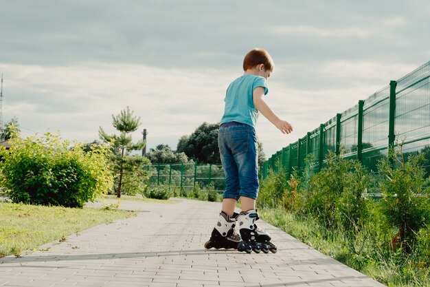 Little boy in protective equipment and rollers stands on walkway in park, low angle view