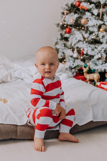 Little boy in red and white pajamas is sitting on the edge of the bed next to the Christmas tree