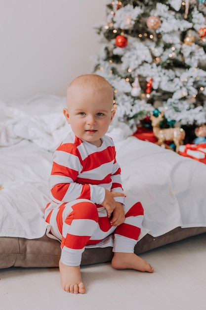 Little boy in red and white pajamas is sitting on the edge of the bed next to the Christmas tree