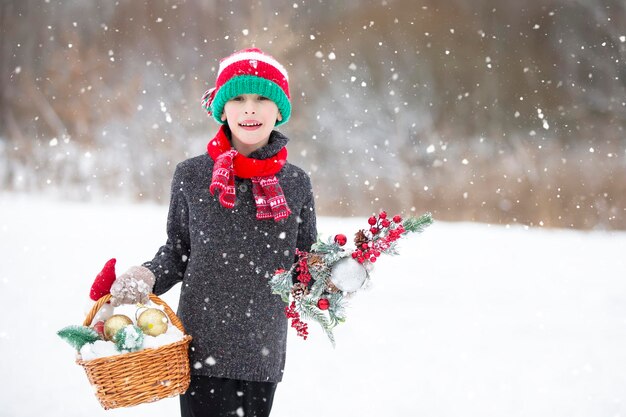 A little boy in a santa claus hat walks through the forest with a christmas decoration