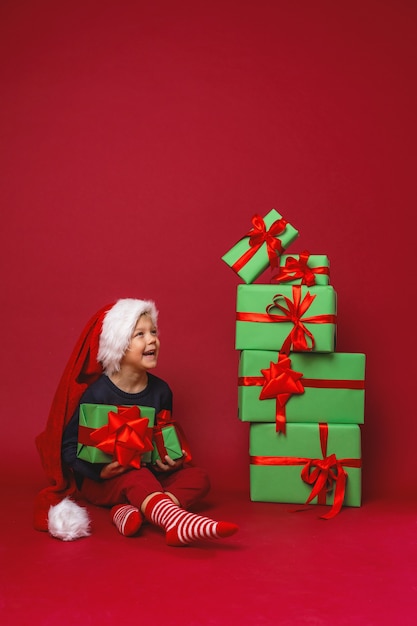 A little boy in a Santa hat sits next to gift boxes