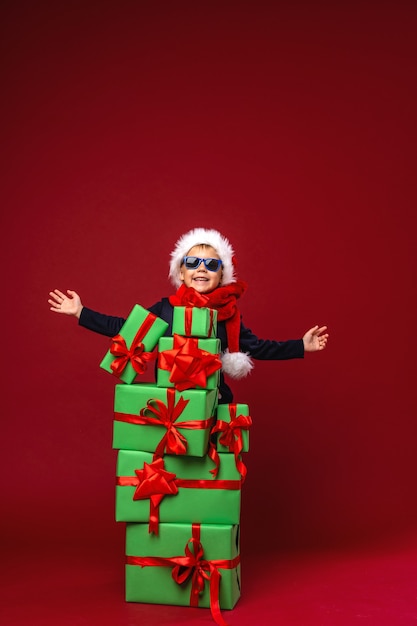 A little boy in a Santa hat stands behind gift boxes