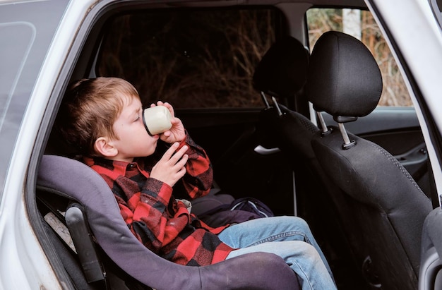 Little boy sitting in a car and drinking from a thermos mug Car seat for children