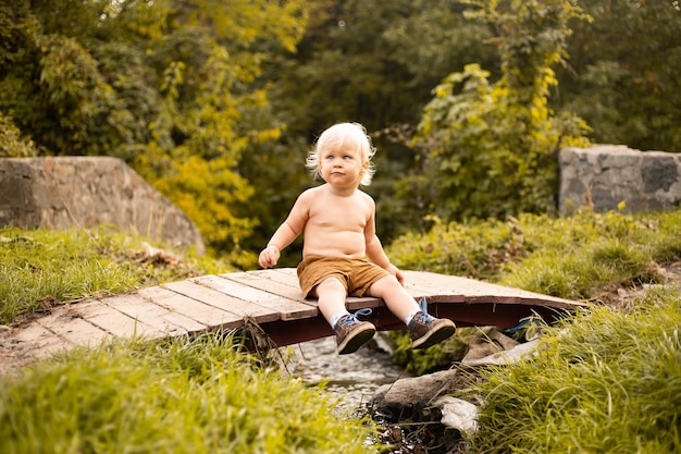 Little boy sitting in shorts on bridge over the stream in autumn or summer park with golden trees