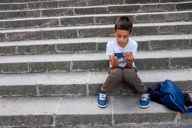 A little a boy sitting with his backpack on stairs with mobile