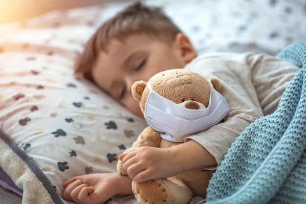 Little boy sleeping in his bed with his Teddy bear wearing a mask to protect him against corona virus covid-19 / 2019-nCov concept.