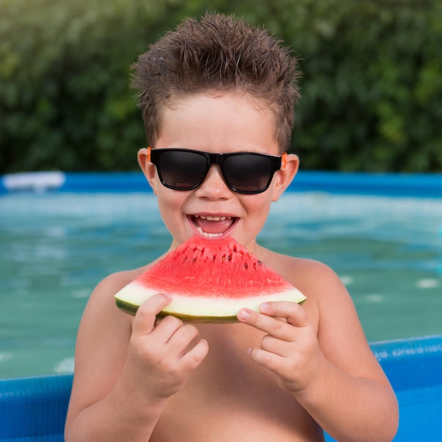 Little boy stands by the pool with a piece of watermelon laughs cheerfully summer vacation