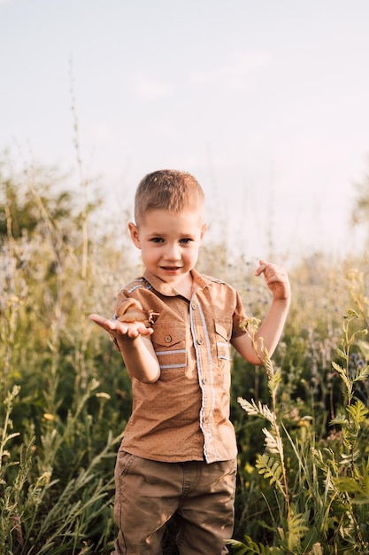 A little boy stands in the grass in nature and holds a snail in his hand