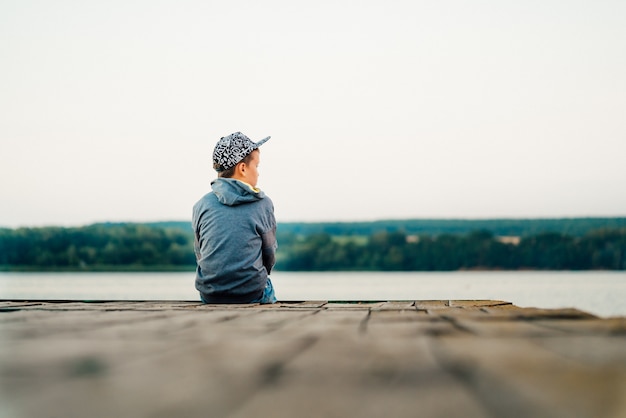 A little boy in a stylish cap and jacket looks out into the distance on the bridge near the lake.