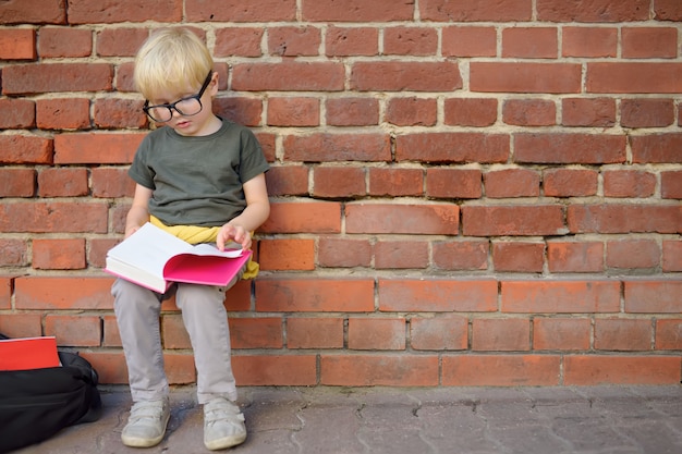 Little boy wearing eyeglass doing homework on break near school building. Back to school concept.