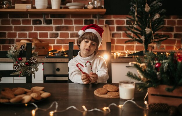 A little boy in a white knitted sweater and a New Year's hat is sitting at the table and holding lollipops on a stick