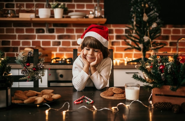 A little boy in a white sweater and a New Year's hat is sitting in the kitchen and dreaming