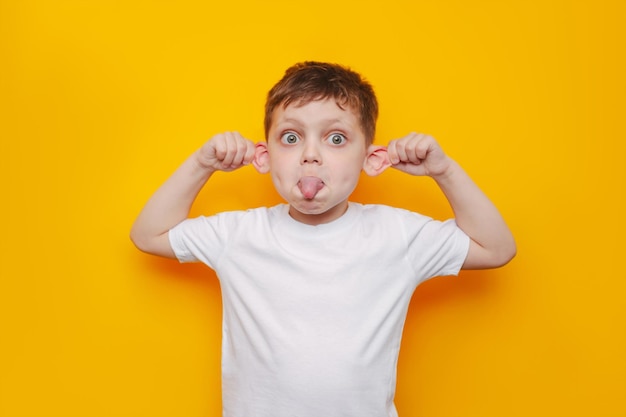 A little boy in a white t-shirt shows tongue pulling the ears. Child grimacing showing funny face