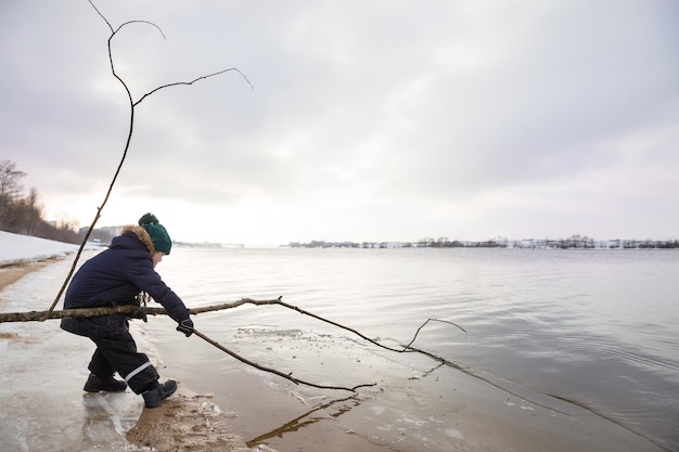 Little boy in winter cloths playing with tree stick and ice on the beach Nature park and outdoors