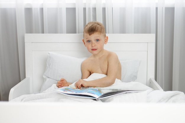 A little boy with blue eyes is reading a book in a white bed