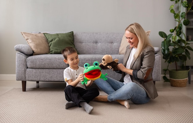 A little boy with his mother is sitting on the carpet near the sofa and playing puppet theater with a monkey and a frog