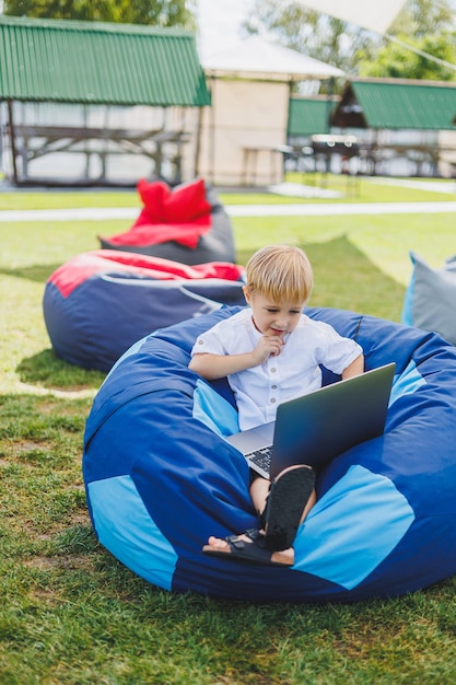 Little boy with a laptop on the background of summer grass A boy in a white Tshirt is sitting on a beanbag in the park