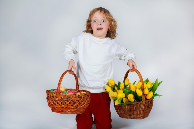 Little boy with long blond curly hair holds in his hands baskets with tulips and easter eggs on white background. Funny moments. Spring holidays concept.