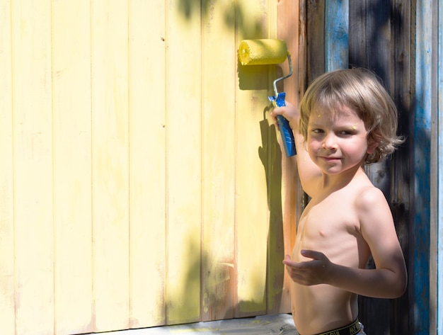 A little boy with a roller paints the house with yellow paint