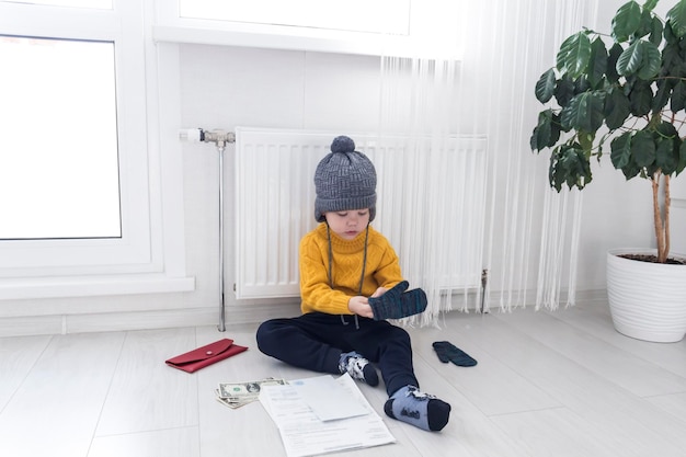 A little boy in a yellow sweater and hat is counting money and studying heating bills near a heater with a thermostat