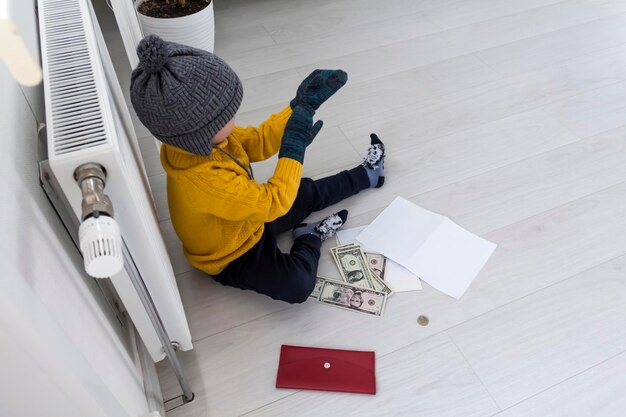 A little boy in a yellow sweater and hat is counting money and studying heating bills near a heater with a thermostat