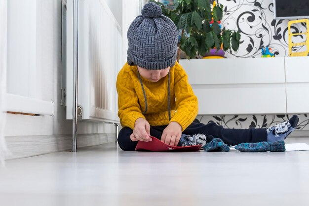 A little boy in a yellow sweater and hat is counting money and studying heating bills near a heater with a thermostat