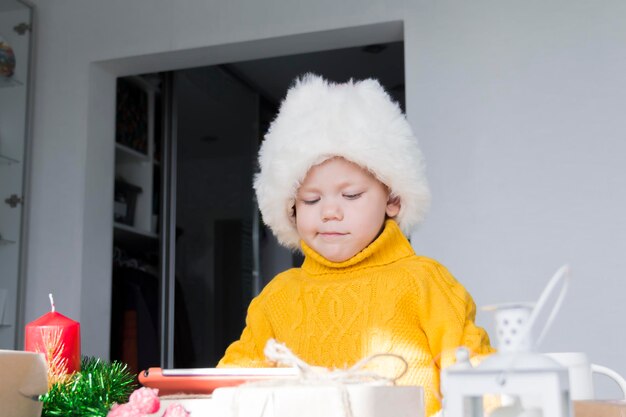 A little boy in a yellow sweater and a red Santa Claus hat at a wooden table with a tablet and in Christmas decorations