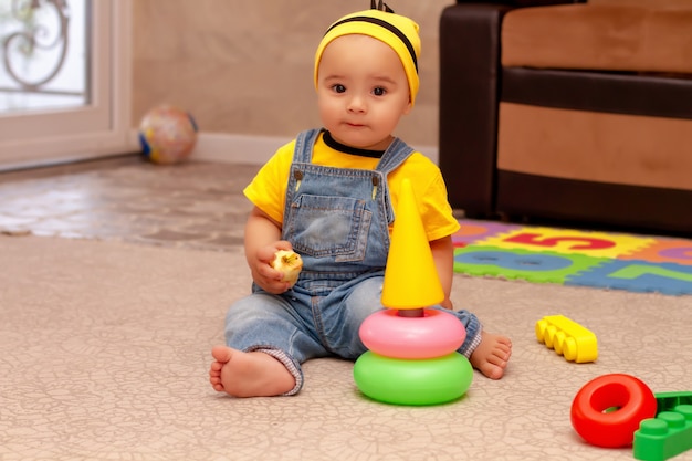 a little boy in a yellow Tshirt a hat and denim overalls is sitting in a room at home