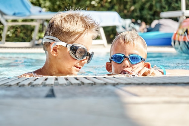 Little boys with swimming goggles have fun in hotel pool water on sunny day at resort close up