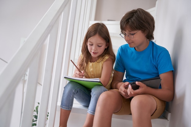 Little brother and sister sitting on stairs at home using digital tablet and learning online or