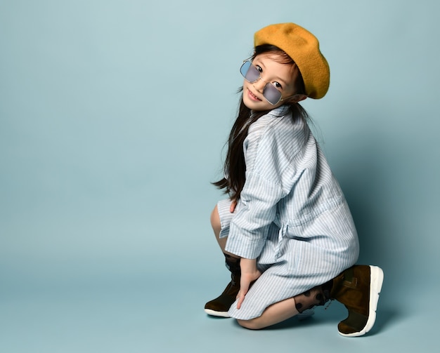 Little brunette asian girl in sunglasses, oversized shirt dress, brown beret, boots. She smiling, posing sitting sideways on blue background. Childhood, fashion, hipster style. Close up, copy space