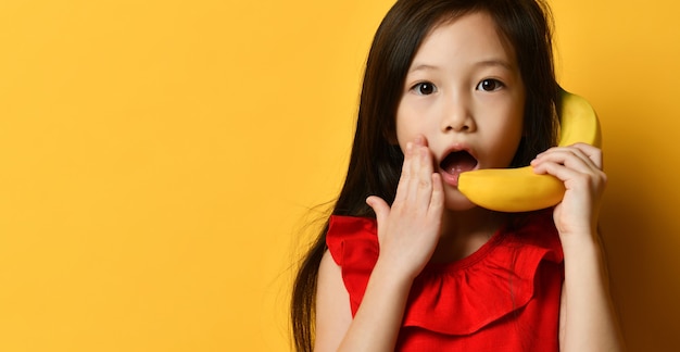 Little brunette asian kid in red blouse. She pretending to be talking on banana like by phone, posing against orange studio background. Childhood, fruits, emotions, advertising. Close up, copy space