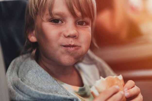 Little candid kid boy five years old eats burger or sandwich food sitting in airplane seat on flight traveling from airport children take a bite child in air plane eating lunch or dinner meal flare