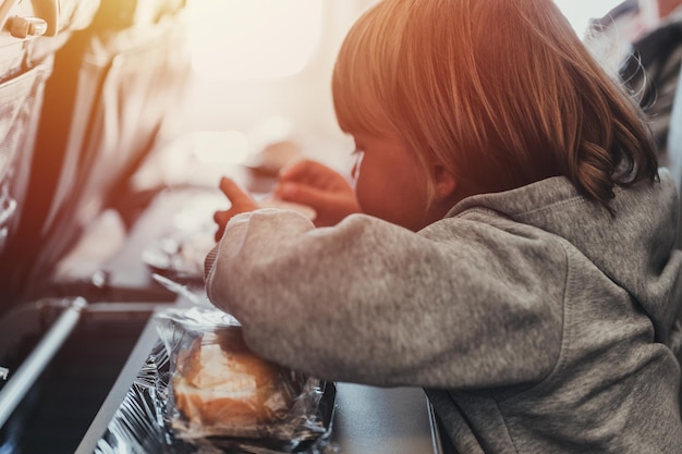 Little candid kid boy five years old eats burger or sandwich food sitting in airplane seat on flight traveling from airport children take a bite child in air plane eating lunch or dinner meal flare