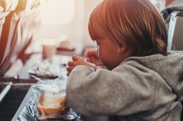 Little candid kid boy five years old eats burger or sandwich food sitting in airplane seat on flight traveling from airport children take a bite child in air plane eating lunch or dinner meal flare