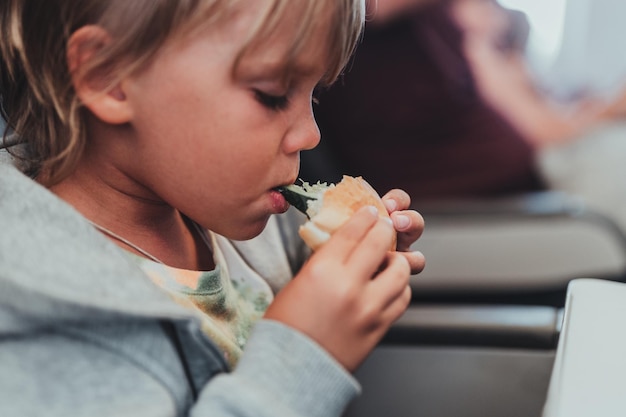 Little candid kid boy five years old eats burger or sandwich food sitting in airplane seat on flight traveling from airport children take a bite child in air plane eating lunch or dinner meal