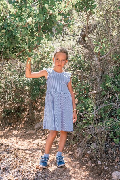 Little candid tired kid girl traveler and hiker of eight years old in a blue striped dress travels and hiking a mountain path among forest green plants in the mediterranean during her summer vacation