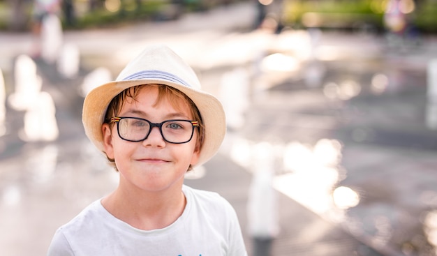 Little caucasian boy in hat and big glasses playing and having fun with water in fountain in the sunny summer park.