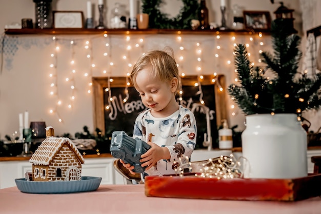 Little caucasian boy playing with a car while standing in the kitchen with new year decor and garlands