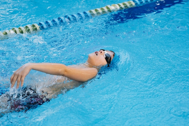 Little caucasian boy wearing goggles swimming backstroke in a pool