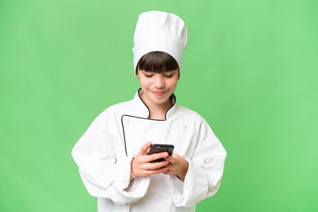 Little caucasian chef girl over isolated background sending a message with the mobile