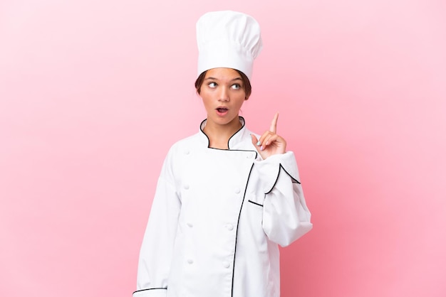 Little caucasian chef girl isolated on pink background thinking an idea pointing the finger up