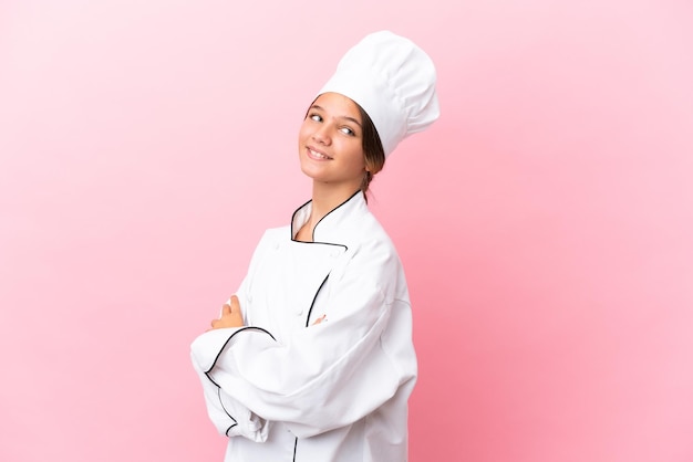 Little caucasian chef girl isolated on pink background with arms crossed and happy