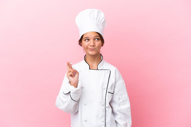 Little caucasian chef girl isolated on pink background with fingers crossing and wishing the best